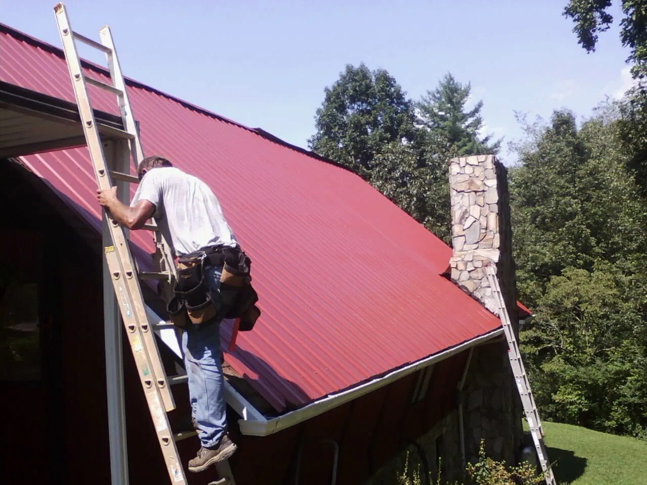 Roofer on a ladder beside a red metal roof and stone chimney — Soffit & Fascia Repair by Roofing Brothers in Rainier